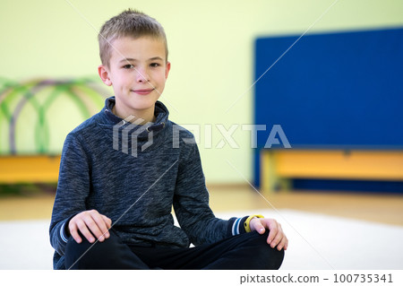 Young child boy sitting and relaxiong on the floor inside sports room in a school after training. 100735341