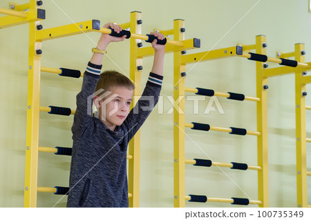 Young child boy exercising on a wall ladder bar inside sports gym room in a school. 100735349