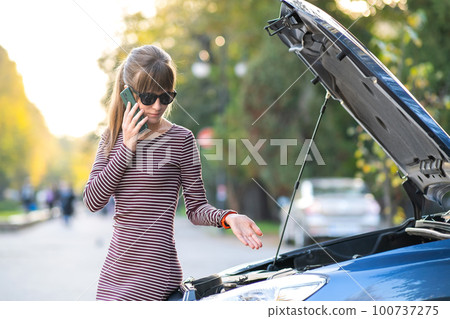 Upset young woman driver talking angrily on cell phone with assistance service near a broken car with open hood on a city street. Upset young woman driver talking angrily on cell phone with assistance service near a broken car with open hood on a city street. 100737275