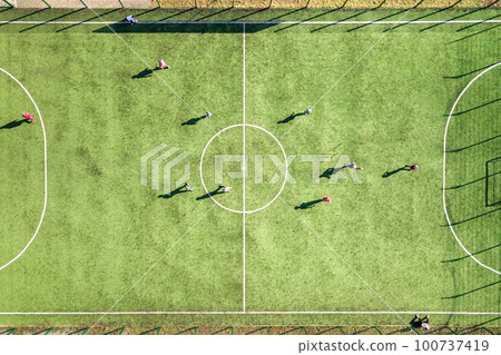 Top down aerial view of green football sports field and players playing football. Drone taken image of small unrecognizable sportsmen on grass covered stadium during sport activities. Top down aerial view of green football sports field and players playing football. Drone taken image of small unrecognizable sportsmen on grass covered stadium during sport activities. 100737419