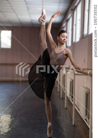 Graceful Asian ballerina in a beige bodysuit and black skirt is rehearsing in a dance class.  100738502