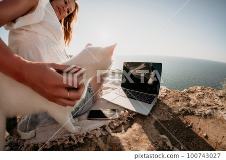 Woman sea laptop. Business woman petting cat and working on laptop by the sea. Close up on hands of pretty lady typing on computer outdoors summer day. Freelance, digital nomad and holidays concept. Woman sea laptop. Business woman petting cat and working on laptop by the sea. Close up on hands of pretty lady typing on computer outdoors summer day. Freelance, digital nomad and holidays concept. 100743027