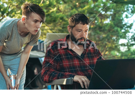 Caucasian bearded man and teenage son working wing with laptop outdoors. Camping concept Caucasian bearded man and teenage son working wing with laptop outdoors. Camping concept 100744508