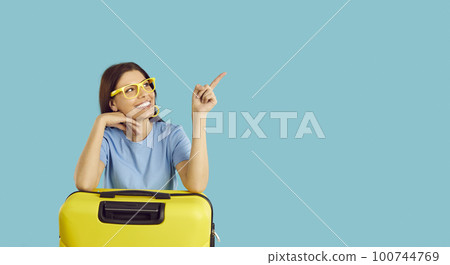 Happy woman with her yellow suitcase smiling and pointing at copy space on blue background 100744769