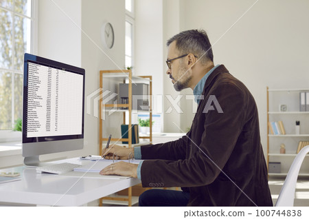 Portrait of serious, attentive, mature businessman man with glasses, sitting at his workplace and looking at his computer screen. 100744838