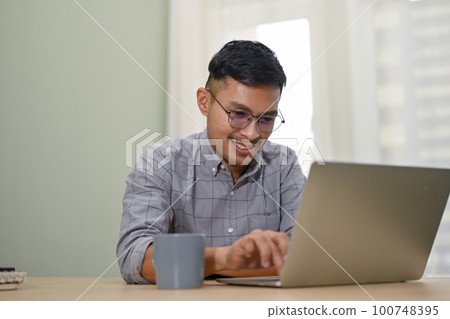 Happy millennial Asian businessman working at his desk in his office, using laptop Happy millennial Asian businessman working at his desk in his office, using laptop 100748395