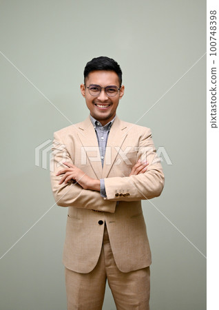 Smart Asian businessman stands against a green studio background with arms crossed. 100748398