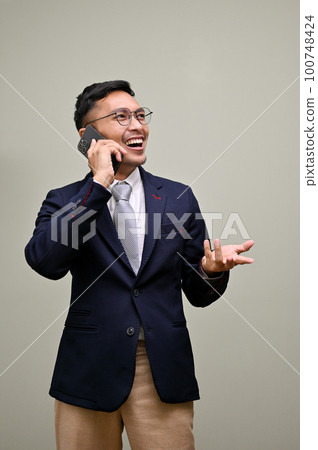 Smiling Asian businessman talking on the phone with someone, standing against a green background. Smiling Asian businessman talking on the phone with someone, standing against a green background. 100748424