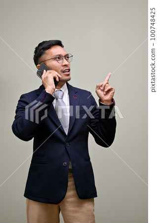 Professional millennial Asian businessman talking on the phone, standing against a green background. 100748425