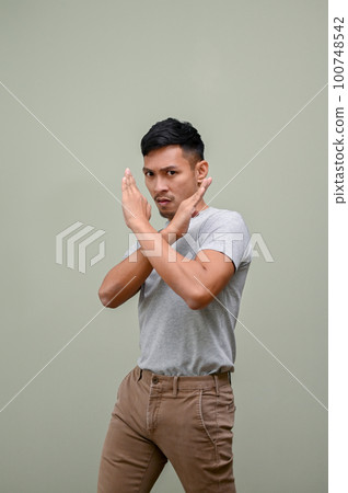 Serious Asian man with crossing hands gesture stands against a green studio background Serious Asian man with crossing hands gesture stands against a green studio background 100748542