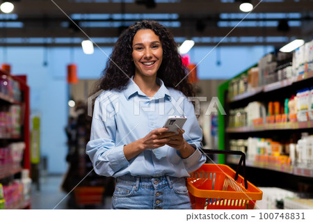 Shopping in a supermarket. A young Latin American woman stands between rows of shelves with a cart. He holds the phone, has a list, writes messages, orders and pays online. Smiling at the camera. Shopping in a supermarket. A young Latin American woman stands between rows of shelves with a cart. He holds the phone, has a list, writes messages, orders and pays online. Smiling at the camera. 100748831