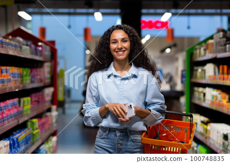 Shopping in a supermarket. A young Hispanic woman walks between rows of shelves with a shopping cart. He holds the phone in his hands, smiles at the camera. Shopping in a supermarket. A young Hispanic woman walks between rows of shelves with a shopping cart. He holds the phone in his hands, smiles at the camera. 100748835