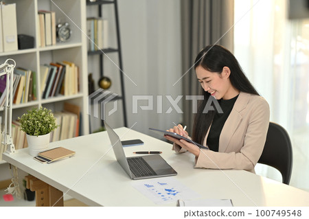 Smiling millennial female manager sitting in front of laptop using digital tablet checking financial data in office 100749548