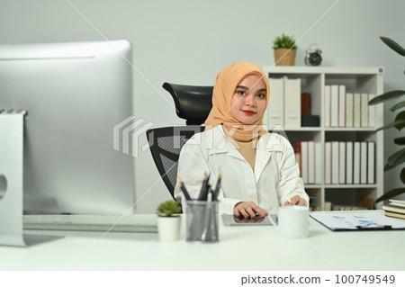 Portrait of confident young working woman in hijab in front of computer monitor at her workplace and smiling to camera 100749549