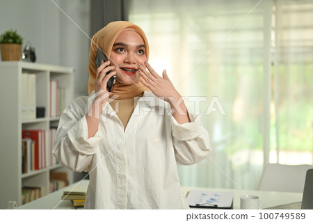 Positive muslim businesswoman consulting client distantly by mobile phone call, standing in modern office 100749589