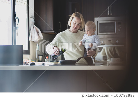 Happy mother and little infant baby boy together making pancakes for breakfast in domestic kitchen. Family, lifestyle, domestic life, food, healthy eating and people concept 100752982