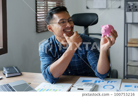 Portrait of an Asian businessman using a calculator to calculate his savings from SME operations, with a pink piggy bank as keep money concept. Portrait of an Asian businessman using a calculator to calculate his savings from SME operations, with a pink piggy bank as keep money concept. 100753216