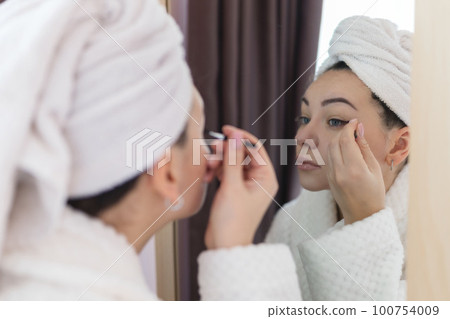 A woman applies black mascara to her eyelashes with a makeup brush. Young beautiful woman applying mascara makeup on her eyes in the bathroom 100754009