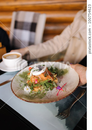Waiter bring breakfast for customer on terrace. Fresh sandwich with salmon and poached egg on top Waiter bring breakfast for customer on terrace. Fresh sandwich with salmon and poached egg on top 100754623