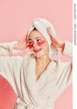 Portrait of smiling happy beautiful girl wearing bathrobe and towel using eye patches for skin refresh over pink background Portrait of smiling happy beautiful girl wearing bathrobe and towel using eye patches for skin refresh over pink background 100756749