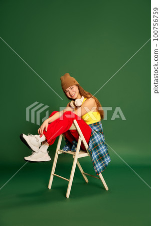 Happy adorable girl wearing stylish clothes, sitting on chair and smiling at camera over green background 100756759