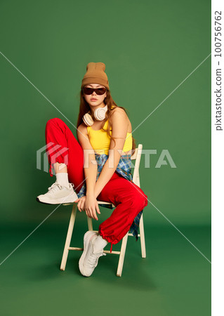 Full-length photo of cool girl wearing stylish clothes, sitting and looking at camera over green background Full-length photo of cool girl wearing stylish clothes, sitting and looking at camera over green background 100756762