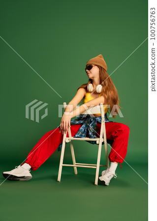 Full-length photo of juvenile girl wearing headphones, sunglasses, sitting on chair and posing over green background 100756763