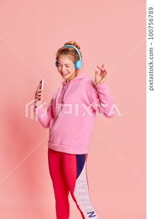 Portrait of dancing young girl wearing headphones and listening music with hands over pink background 100756789