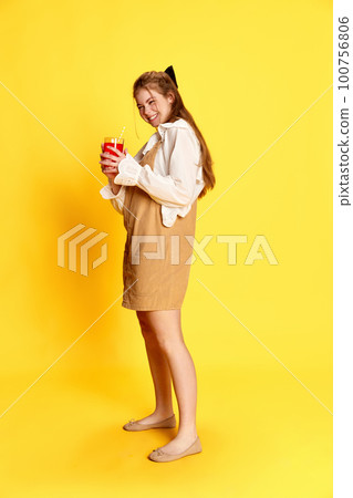 Portrait of young redhaired girl wearing white blouse and dress holding cup of drink and smiling over yellow background Portrait of young redhaired girl wearing white blouse and dress holding cup of drink and smiling over yellow background 100756806