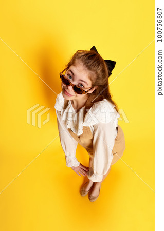 Portrait of young beautiful girl with long hair wearing bow and sunglasses posing at camera like doll over yellow background. Fish eye effect 100756807