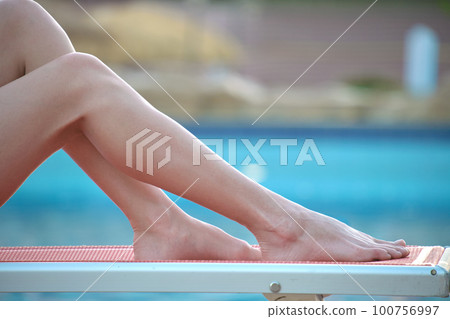 Slim female legs in short white dress relaxing near swimming pool lying down on deck chair. Summer vacation concept Slim female legs in short white dress relaxing near swimming pool lying down on deck chair. Summer vacation concept 100756997