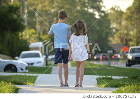 Rear view of two young teenage children, girl and boy, brother and sister walking together on rural street on bright sunny day. Vacation time concept Rear view of two young teenage children, girl and boy, brother and sister walking together on rural street on bright sunny day. Vacation time concept 100757172