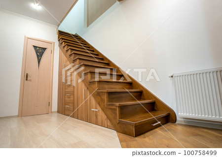 Modern architecture interior with luxury hallway with glossy wooden stairs in multi-storey house. Custom built pullout cabinets on glides in slots under stairs. Use of space for storage. Modern architecture interior with luxury hallway with glossy wooden stairs in multi-storey house. Custom built pullout cabinets on glides in slots under stairs. Use of space for storage. 100758499