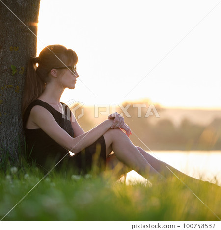 Lonely young woman sitting alone on grass lawn on lake shore enjoying warm evening. Wellbeing and relaxing in nature concept 100758532