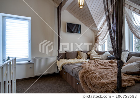 Interior of a modern house spacious hallway with big soft resting place. Contemporary wide sofa with many pillows and light window onder wooden loft ceiling. 100758588