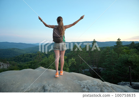 Hiker woman standing on mountain footpath raising up hands enjoying evening nature during travel on wilderness trail. Lonely female traveler traversing high hilltop route. Healthy lifestyle concept Hiker woman standing on mountain footpath raising up hands enjoying evening nature during travel on wilderness trail. Lonely female traveler traversing high hilltop route. Healthy lifestyle concept 100758635