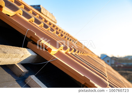 Closeup of metal montage anchor for installation of yellow ceramic roofing tiles mounted on wooden boards covering residential building roof under construction. Closeup of metal montage anchor for installation of yellow ceramic roofing tiles mounted on wooden boards covering residential building roof under construction. 100760723