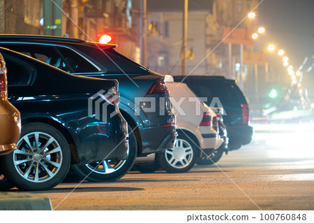 Close up of parked cars on roadside at night with blurred view of traffic lights of moving vehicles on city street. Close up of parked cars on roadside at night with blurred view of traffic lights of moving vehicles on city street. 100760848