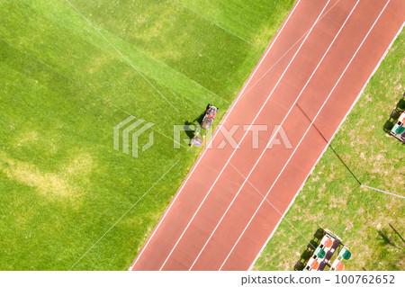 Aerial view of small figure of worker cutting green grass with mowing mashine on football stadium field with red running tracks in summer. 100762652