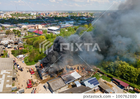 Aerial view of firefighters extinguishing ruined building on fire with collapsed roof and rising dark smoke. 100762880