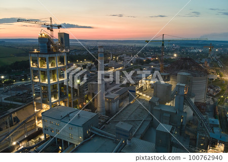 Aerial view of cement factory with high concrete plant structure and tower cranes at industrial production area at night. Manufacture and global industry concept. 100762940