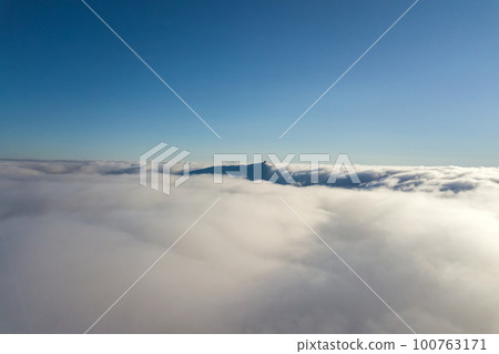 Aerial view from above of white puffy clouds and distant mountain top in bright sunny day. 100763171