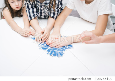 Cropped unrecognizable children hands hold blue playing cards on white table. Smart game for time, who first. Family fun 100763190