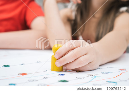 Close up child hand with yellow game paw figure on creative art map. Girl and other kids playing board game with pieces Close up child hand with yellow game paw figure on creative art map. Girl and other kids playing board game with pieces 100763242