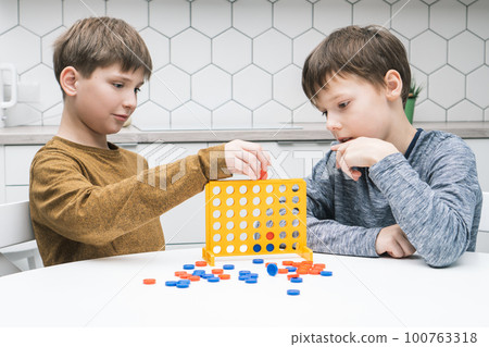 Two positive pensive, concentrated little boys play connect four game with yellow plastic field with holes for figures Two positive pensive, concentrated little boys play connect four game with yellow plastic field with holes for figures 100763318