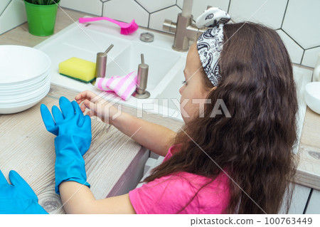 Little girl taking off blue household gloves after washing up dish. Child washing tableware in kitchen sink side view. 100763449