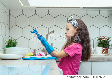 Nice little girl putting on blue household gloves for washing up dish. Home cleaning, personal protective equipment. 100763460