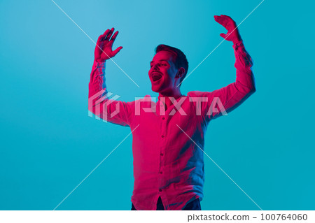 Portrait of young emotional man raising hands with positive happy expression, posing against blue studio background in pink neon light. 100764060