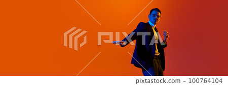 Portrait of young stylish man in jacket and glasses posing, dancing against orange studio background in neon light. 100764104