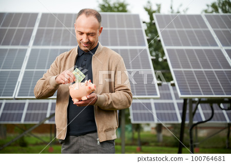 Confidence man putting earned money to piggy bank. Male adult saving profit he got from investing in green energy. Man in casual clothes holding moneybox and putting euros in it. Confidence man putting earned money to piggy bank. Male adult saving profit he got from investing in green energy. Man in casual clothes holding moneybox and putting euros in it. 100764681